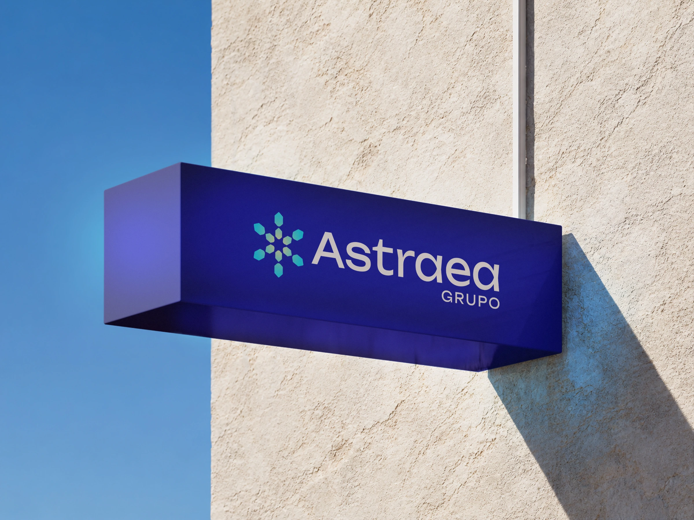 Letrero corpóreo azul con el logotipo de Astraea Grupo en una fachada beige bajo un cielo azul, identidad visual por Mairu Studio