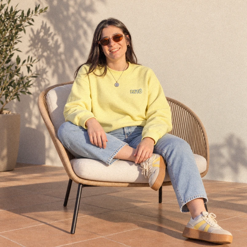 Marina sentada en una butaca de mimbre con sudadera amarilla y gafas de sol